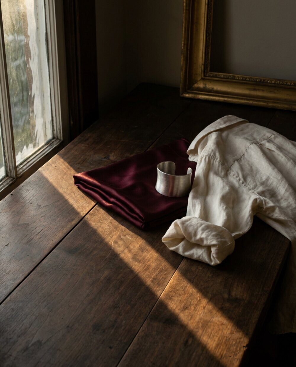 Bodegón sobre una mesa de madera oscura junto a una ventana: una seda burdeos doblada, una camisa de lino crema arrugada con una manga enrollada, y un brazalete ancho de plata mate apoyado encima. Luz baja de tarde entrando por la izquierda, con una sombra diagonal dura cruzando la mesa. Al fondo, el ángulo de un marco dorado viejo.