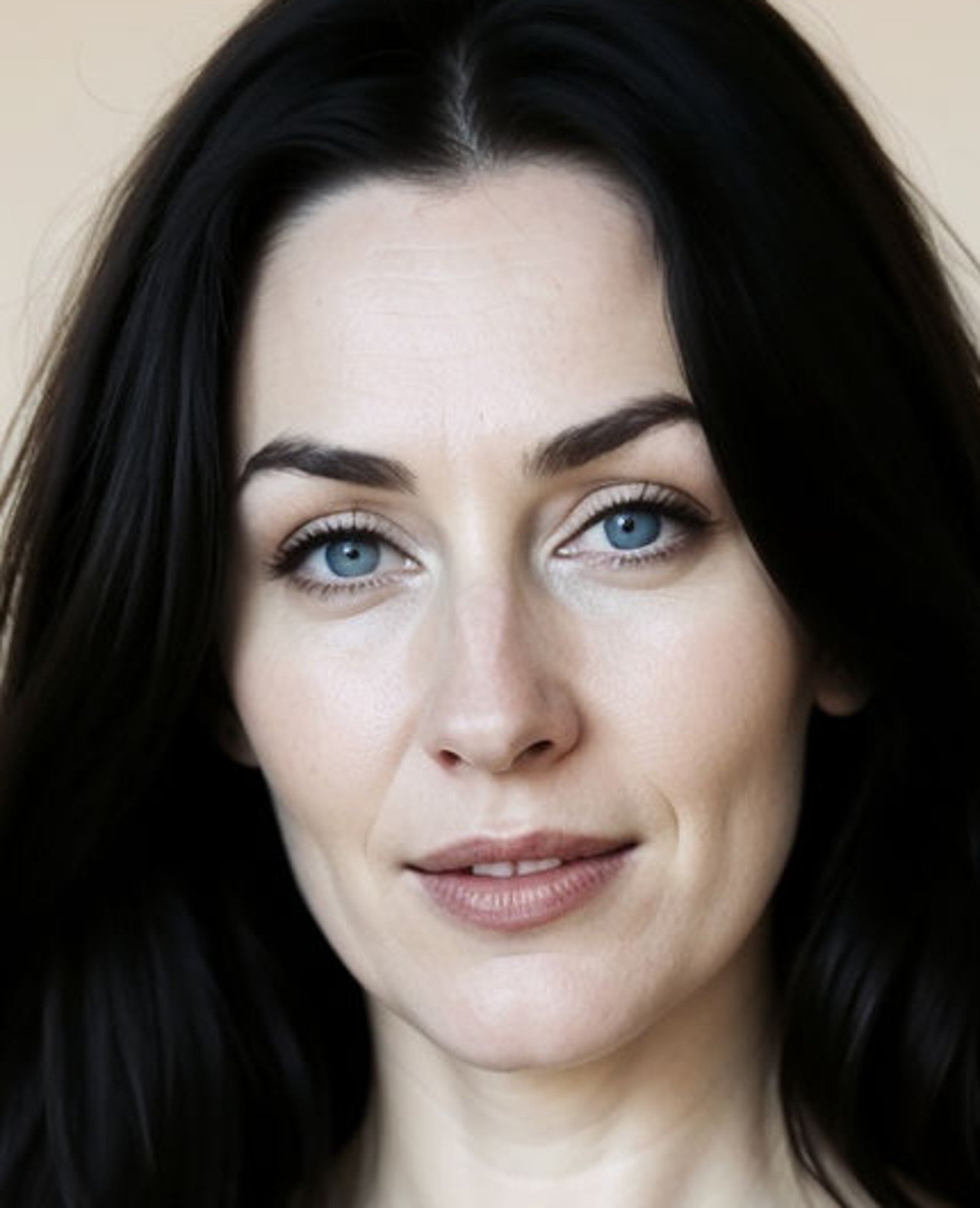 Recorte del rostro de Valentina Serrano ampliado al 300 %: mujer de pelo negro y ojos azules sobre fondo crema, con la textura de la piel visible — poros en la mejilla, líneas finas en la frente, velillo sobre el labio superior y la piel irregular bajo los ojos.